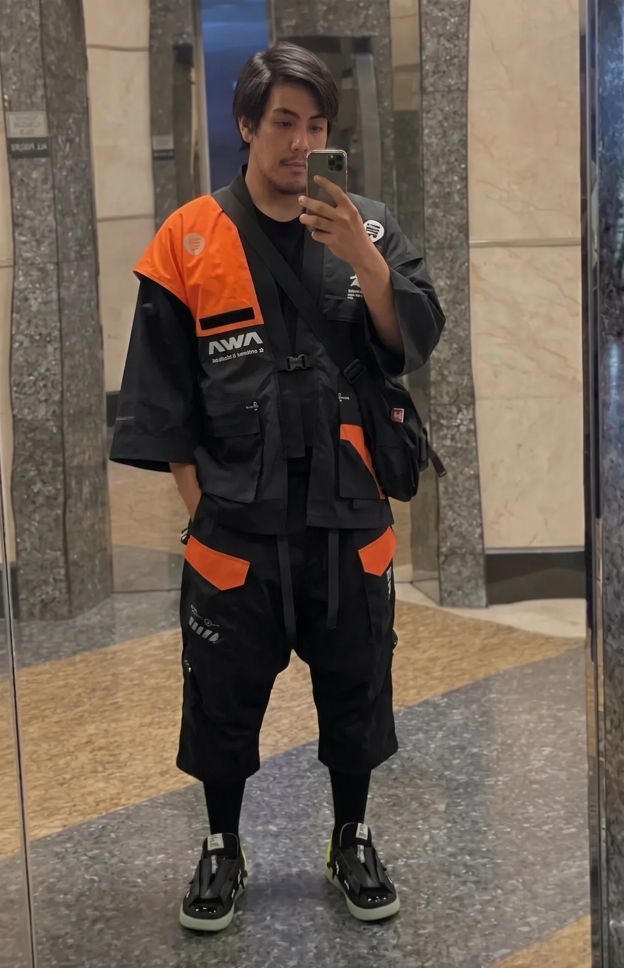 Nicco in a black and orange techwear vest and cargo shorts, mirror selfie